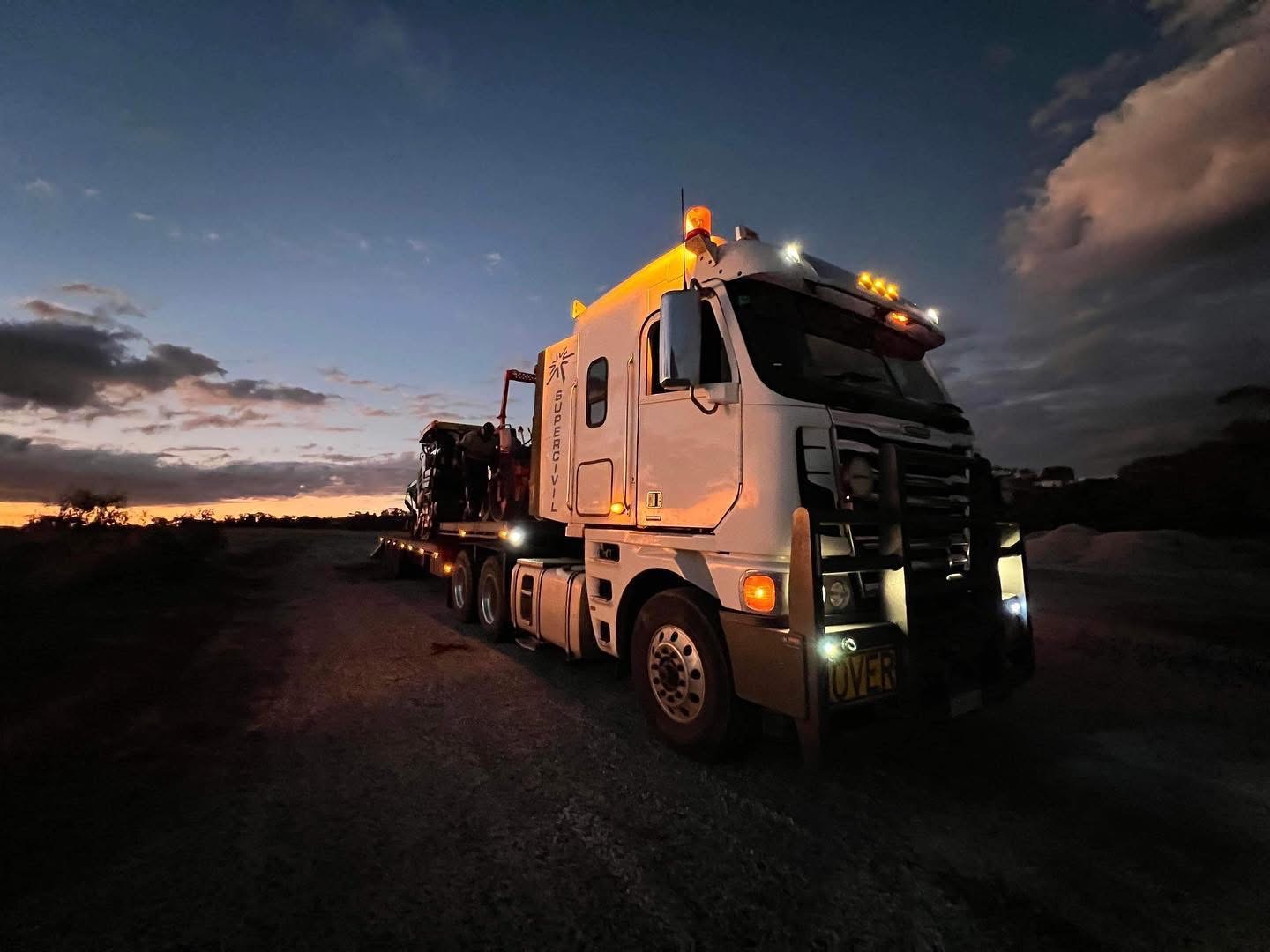SuperCivil Freightliner prime mover hauling heavy plant on a low loader along a rural Western Australian haul road at dusk