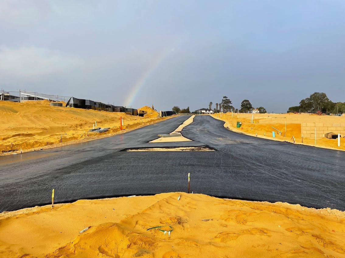 Freshly sealed road through a new Western Australian residential subdivision with sand pads, homes under construction and a rainbow overhead